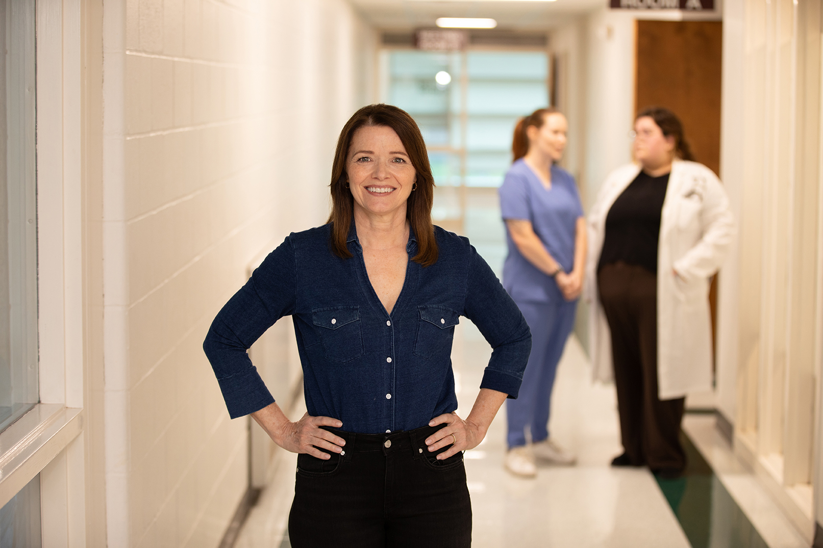 Christina Bohannan in a denim shirt standing with hands on her hips in a hospital hallway, two staff members talking behind her