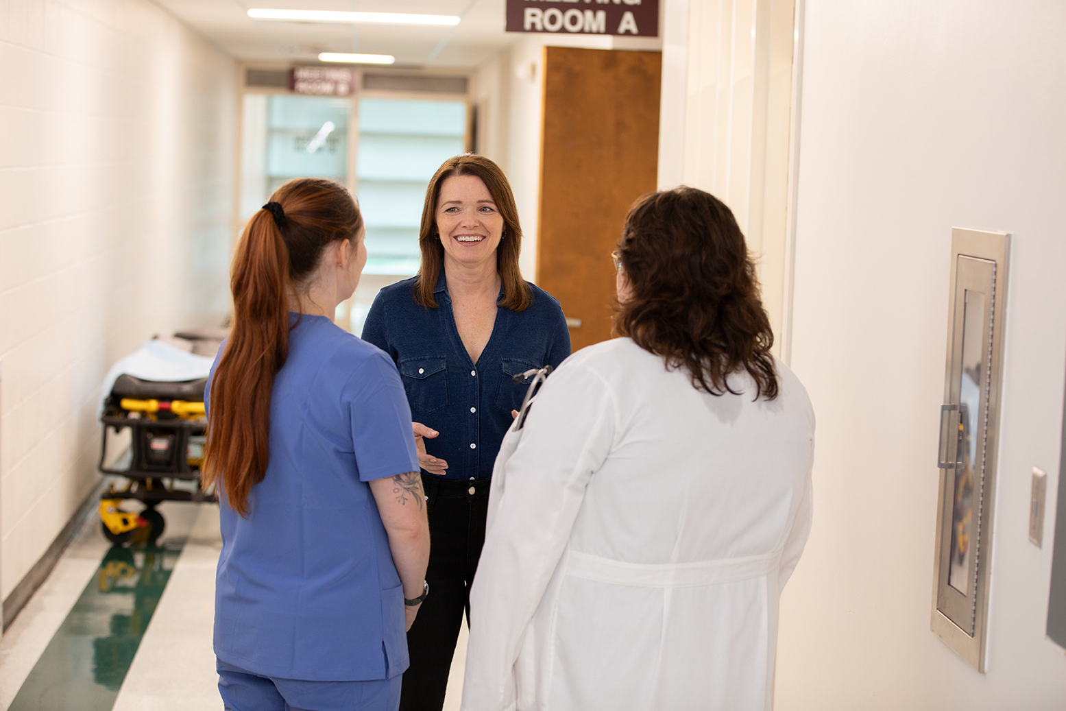 Christina Bohannan in a denim shirt smiling and talking with a nurse in scrubs and a person in a white lab coat in a hospital hallway