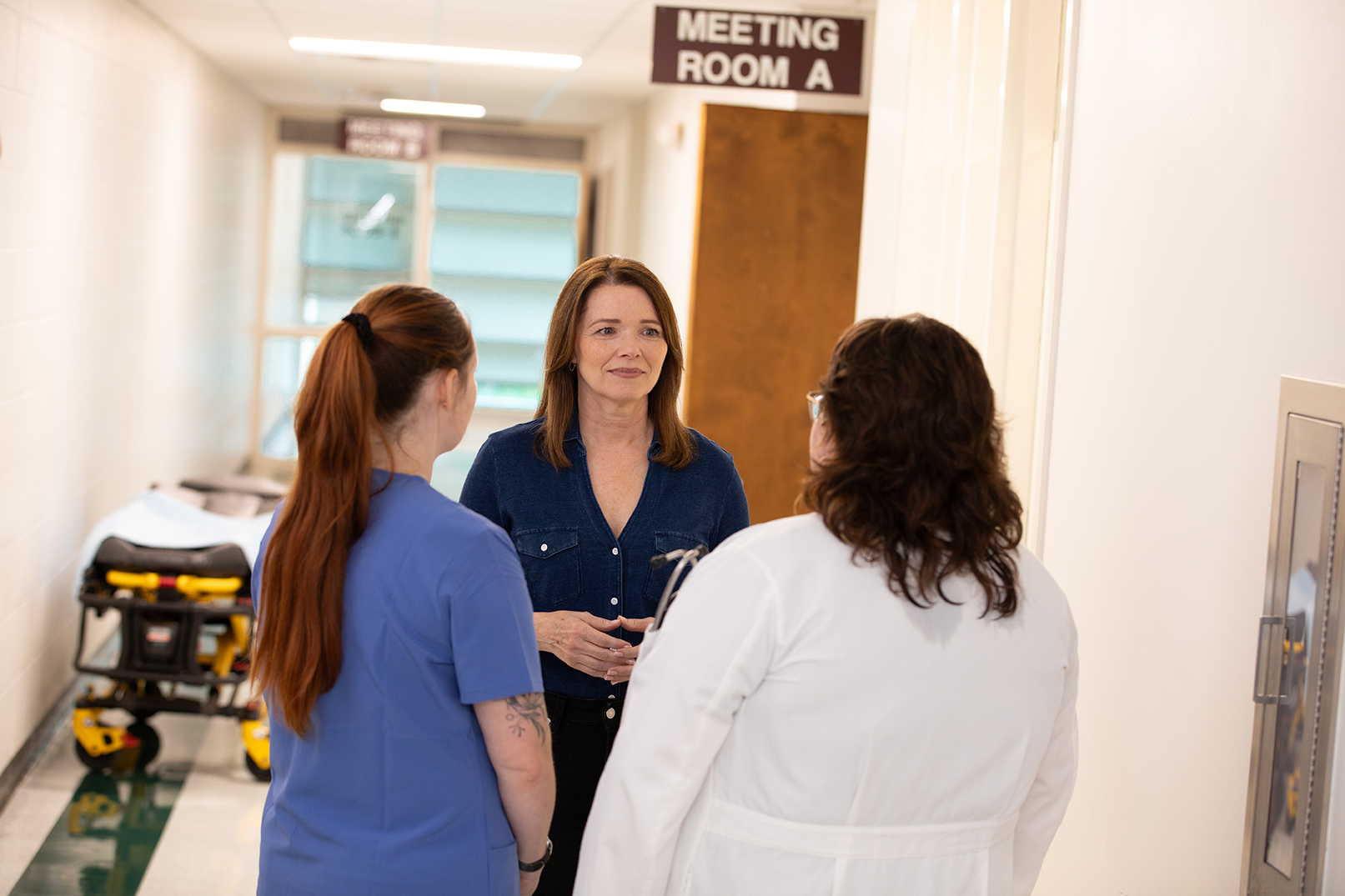 Christina Bohannan in a denim shirt talking with a nurse in blue scrubs and a person in a white coat in a hospital hallway near a Meeting Room A sign