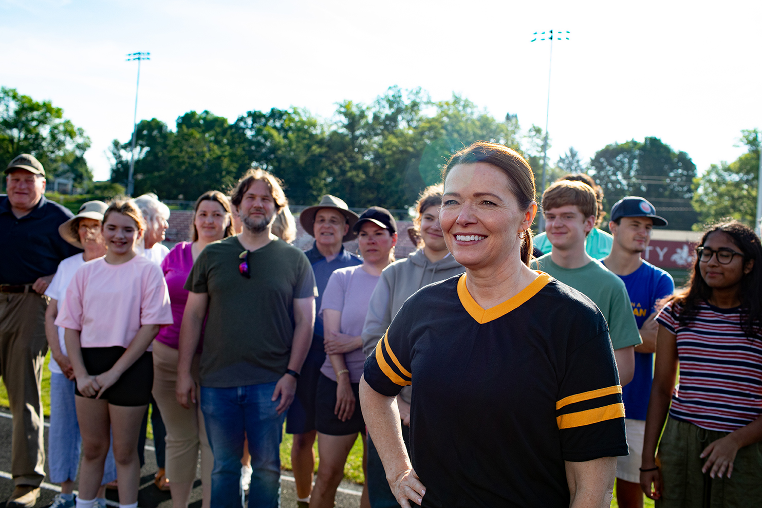 Christina Bohannan in a black and gold jersey smiling, standing in front of a group of people gathered outdoors on a track