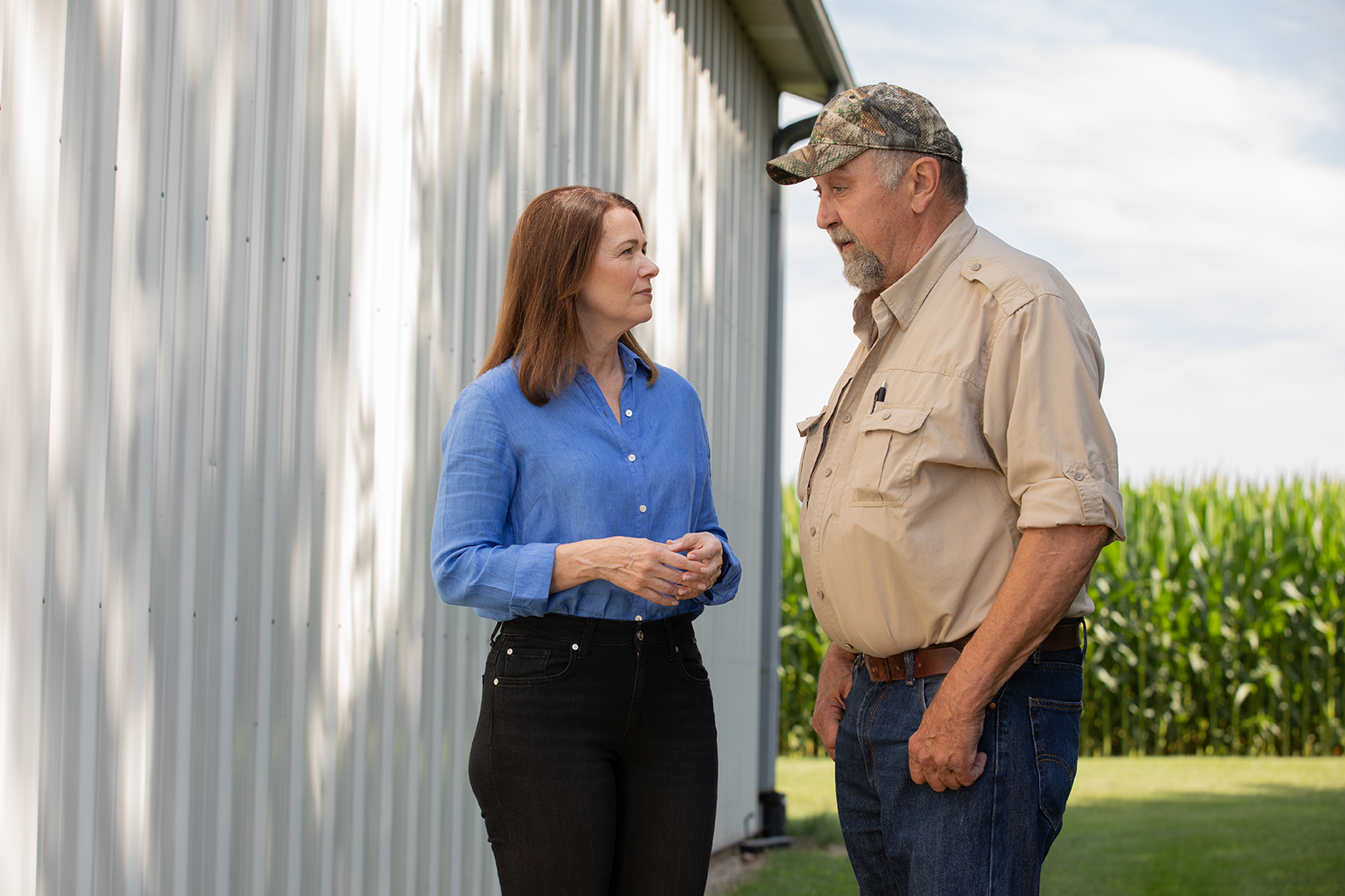 Christina Bohannan in a blue shirt talking with an older man in a camouflage cap and tan shirt beside a metal barn, cornfield in the background