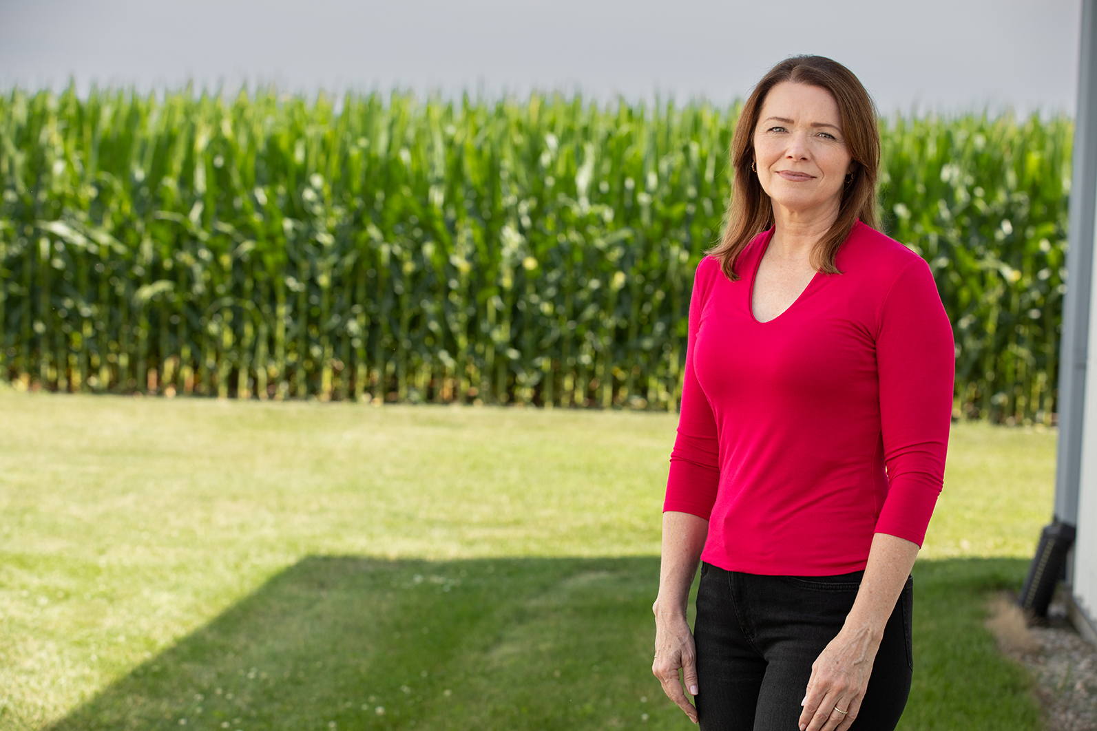 Christina Bohannan in a red top standing in front of a green cornfield on a sunny day