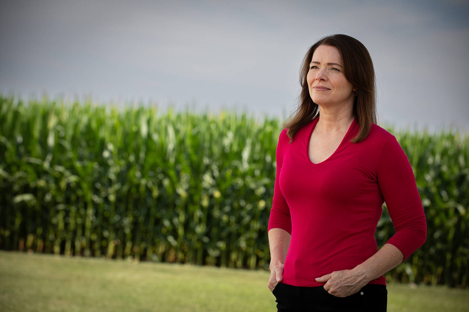 Christina Bohannan in a red top standing in front of a green cornfield, looking into the distance