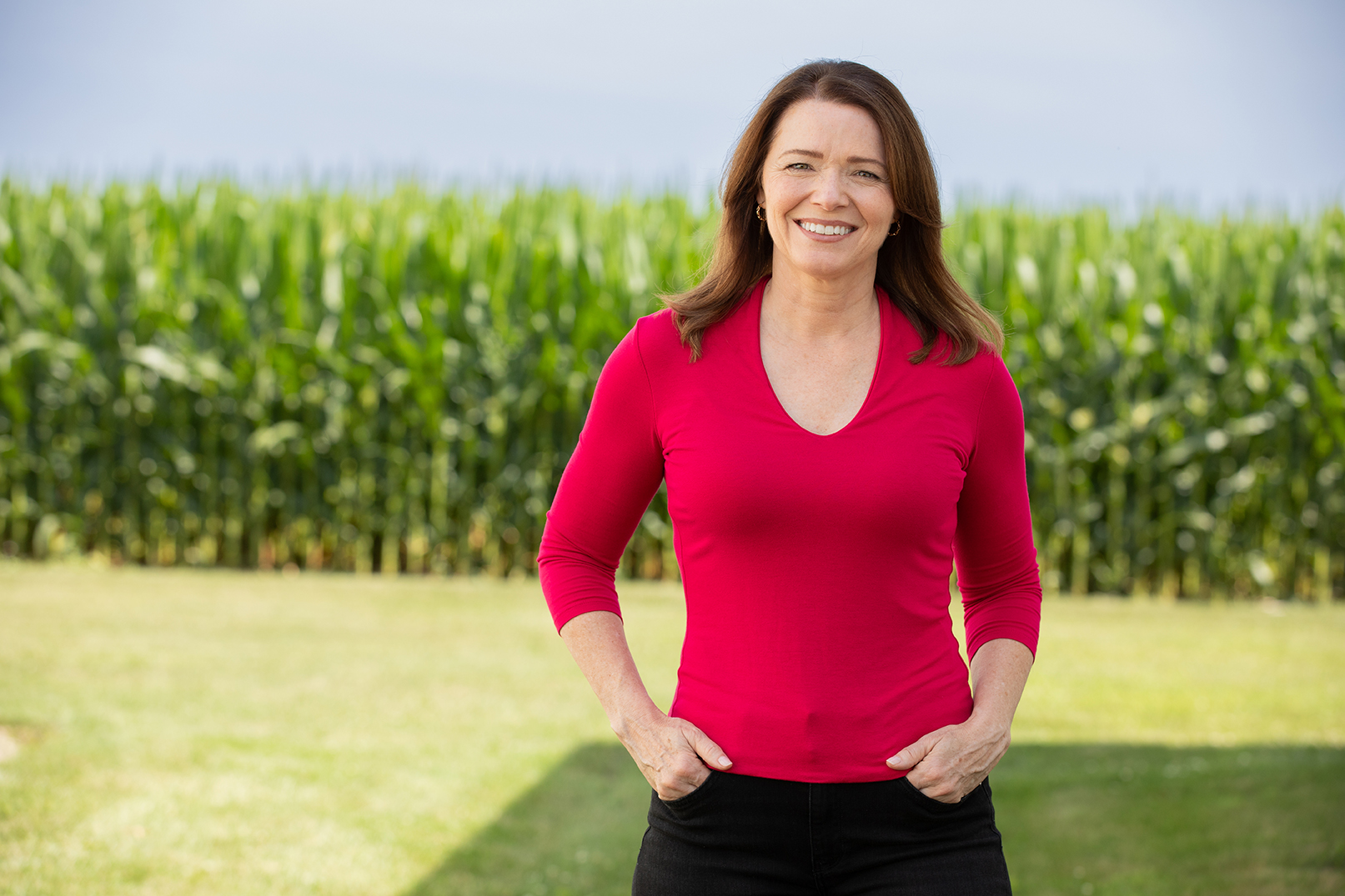 Christina Bohannan in a red top standing with hands in her pockets, smiling, in front of a green cornfield