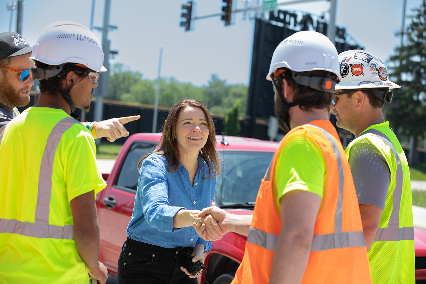 Christina Bohannan in a blue shirt shaking hands with a construction worker in a hard hat and safety vest, other workers around them near a red truck
