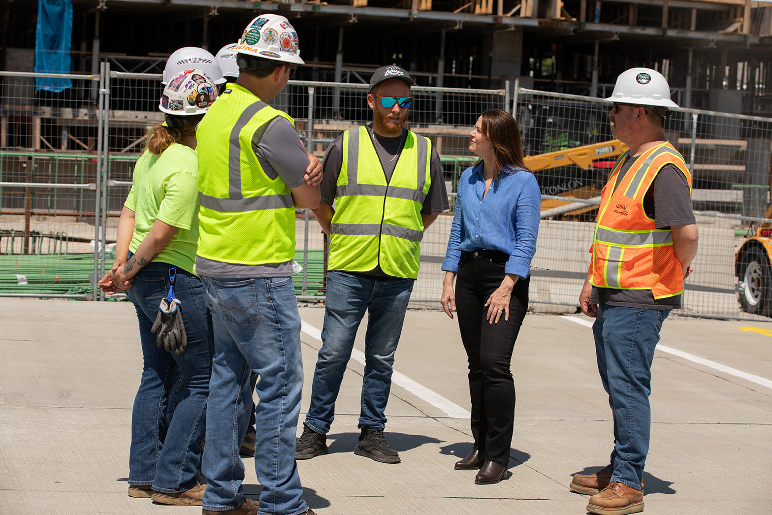 Christina Bohannan in a blue shirt talking with a group of construction workers in hard hats and safety vests at a fenced construction site