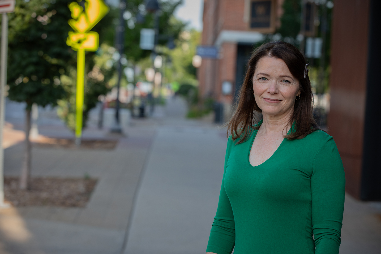Christina Bohannan in a green top standing on a downtown sidewalk with shops and a yield sign in the background
