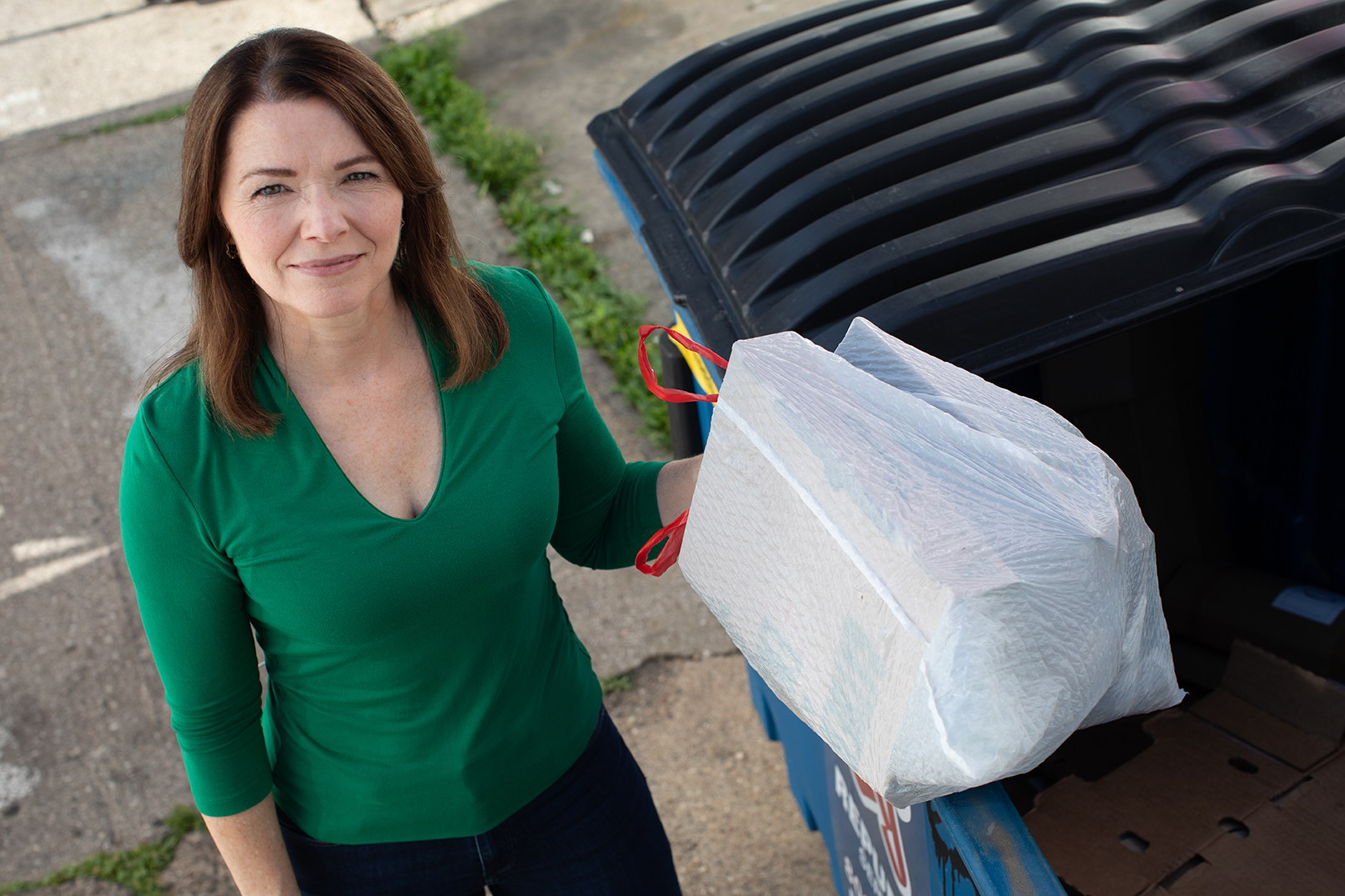 Christina Bohannan in a green top holding a white trash bag beside a blue dumpster outdoors, viewed from above