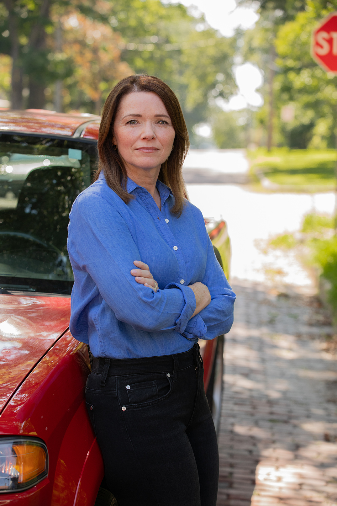 Christina Bohannan in a blue button-up shirt with arms crossed, leaning against a red truck near a stop sign on a brick street