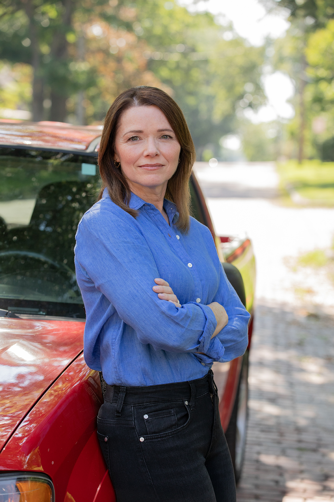 Christina Bohannan in a blue button-up shirt with arms crossed, leaning against a red pickup truck on a residential street