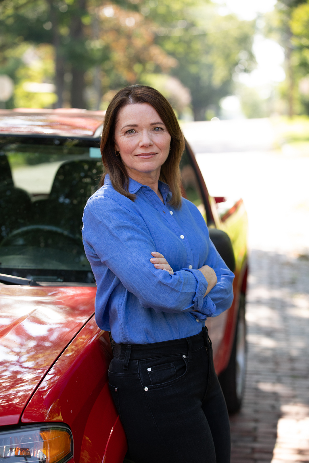 Christina Bohannan in a blue button-up shirt and black jeans with arms crossed, leaning against a red truck on a brick street