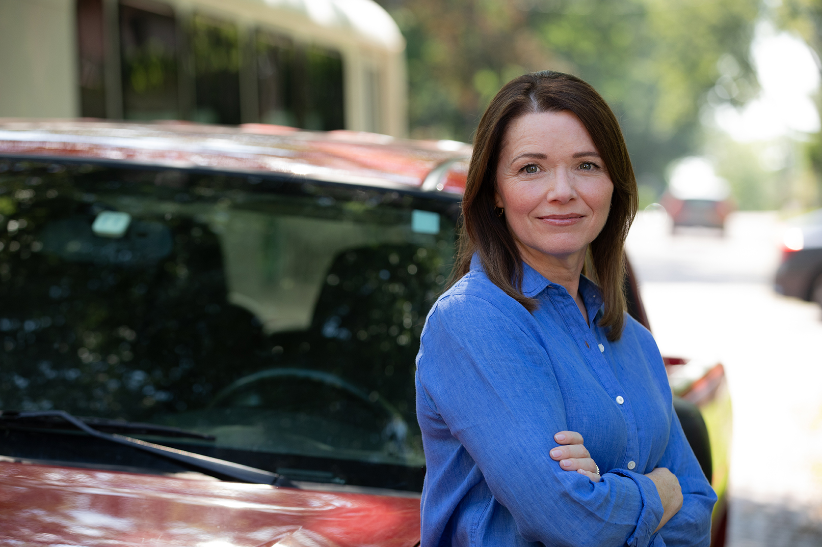 Christina Bohannan in a blue button-up shirt with arms crossed, smiling slightly, standing beside a dark red vehicle