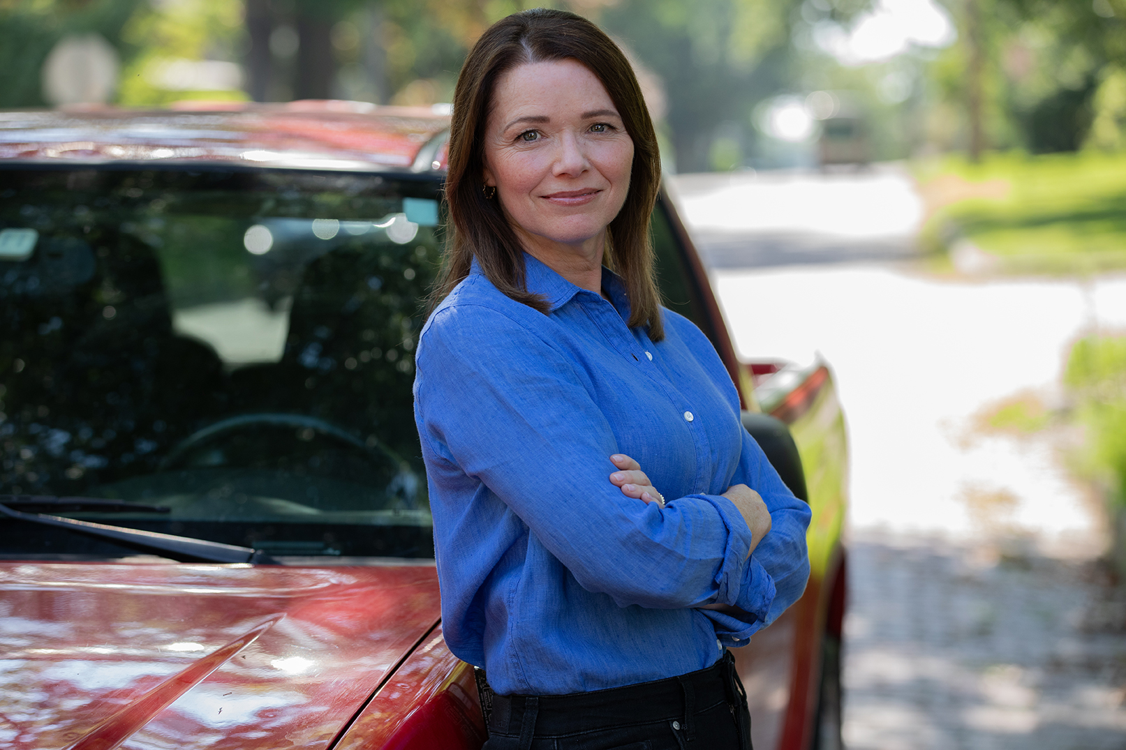 Christina Bohannan in a blue button-up shirt with arms crossed, leaning against a red pickup truck on a tree-lined street