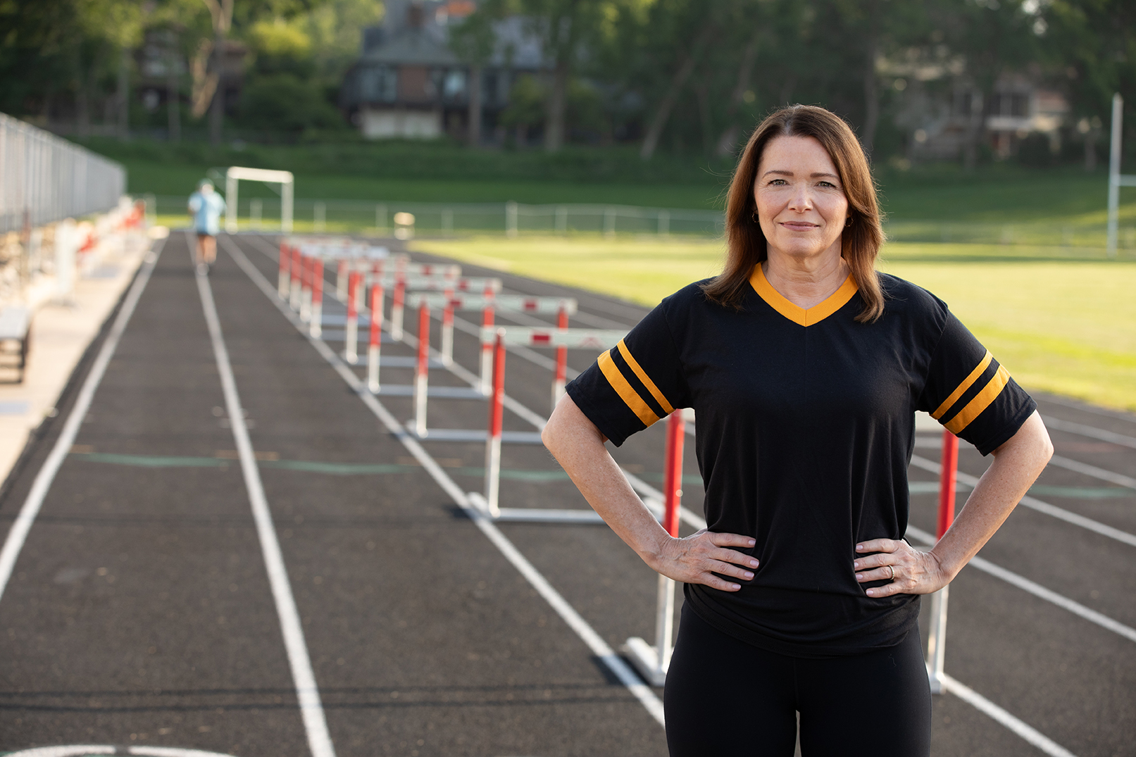 Christina Bohannan in a black and gold jersey standing with hands on hips on a running track lined with hurdles