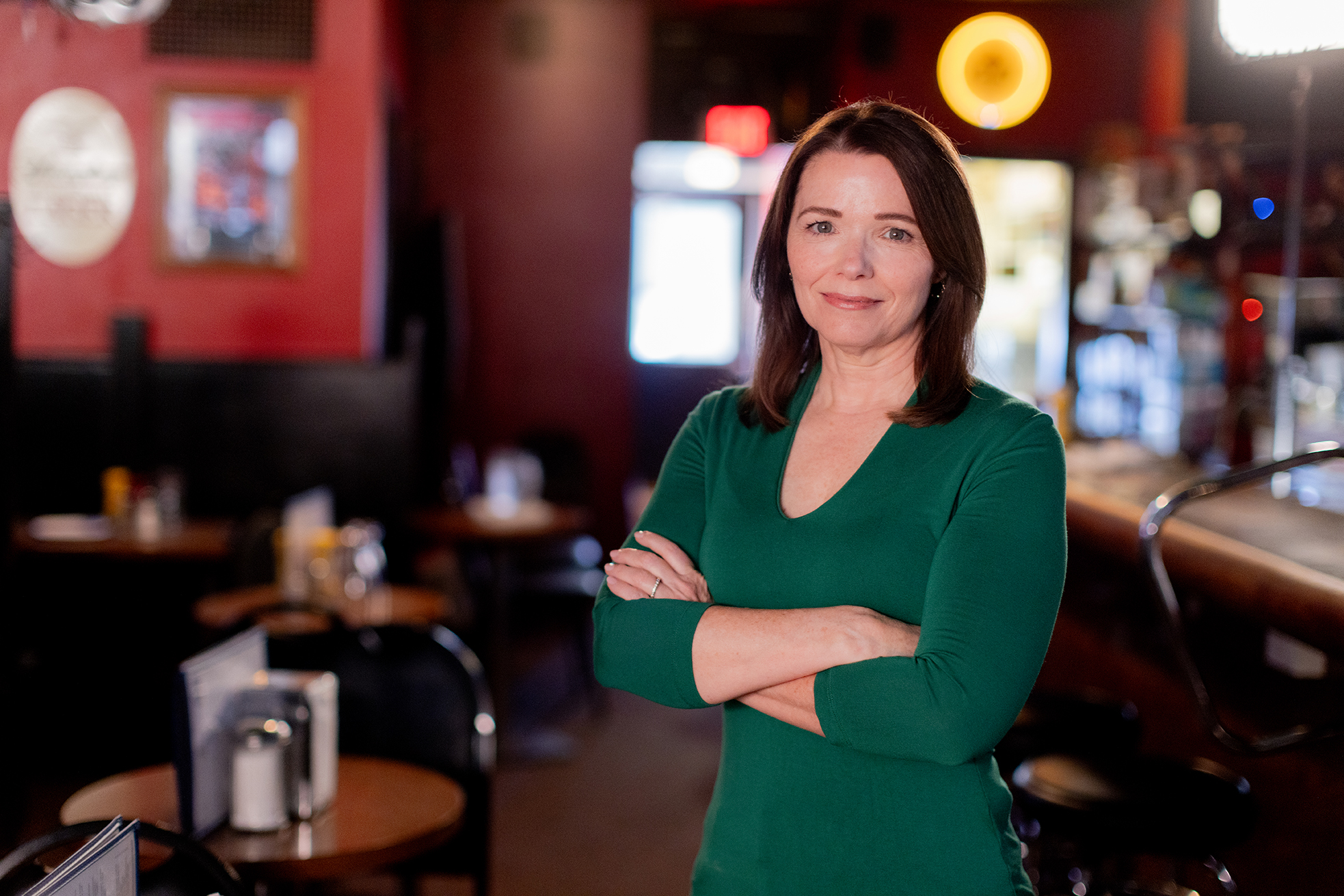 Christina Bohannan in a green top standing with arms crossed inside a dimly lit bar with tables in the background