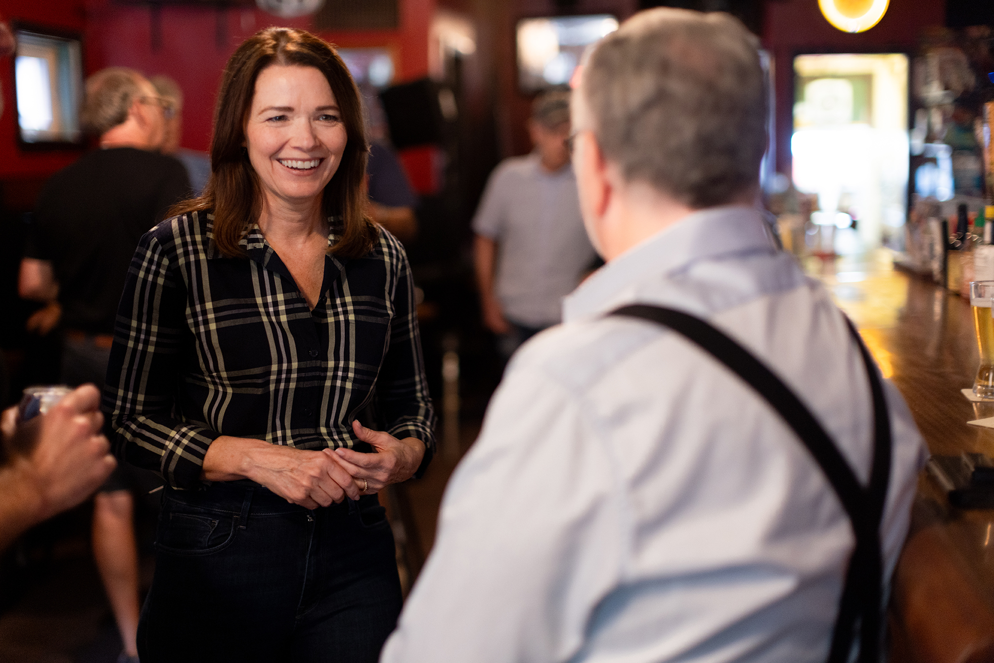 Christina Bohannan in a plaid shirt smiling while talking with a man in suspenders inside a busy bar