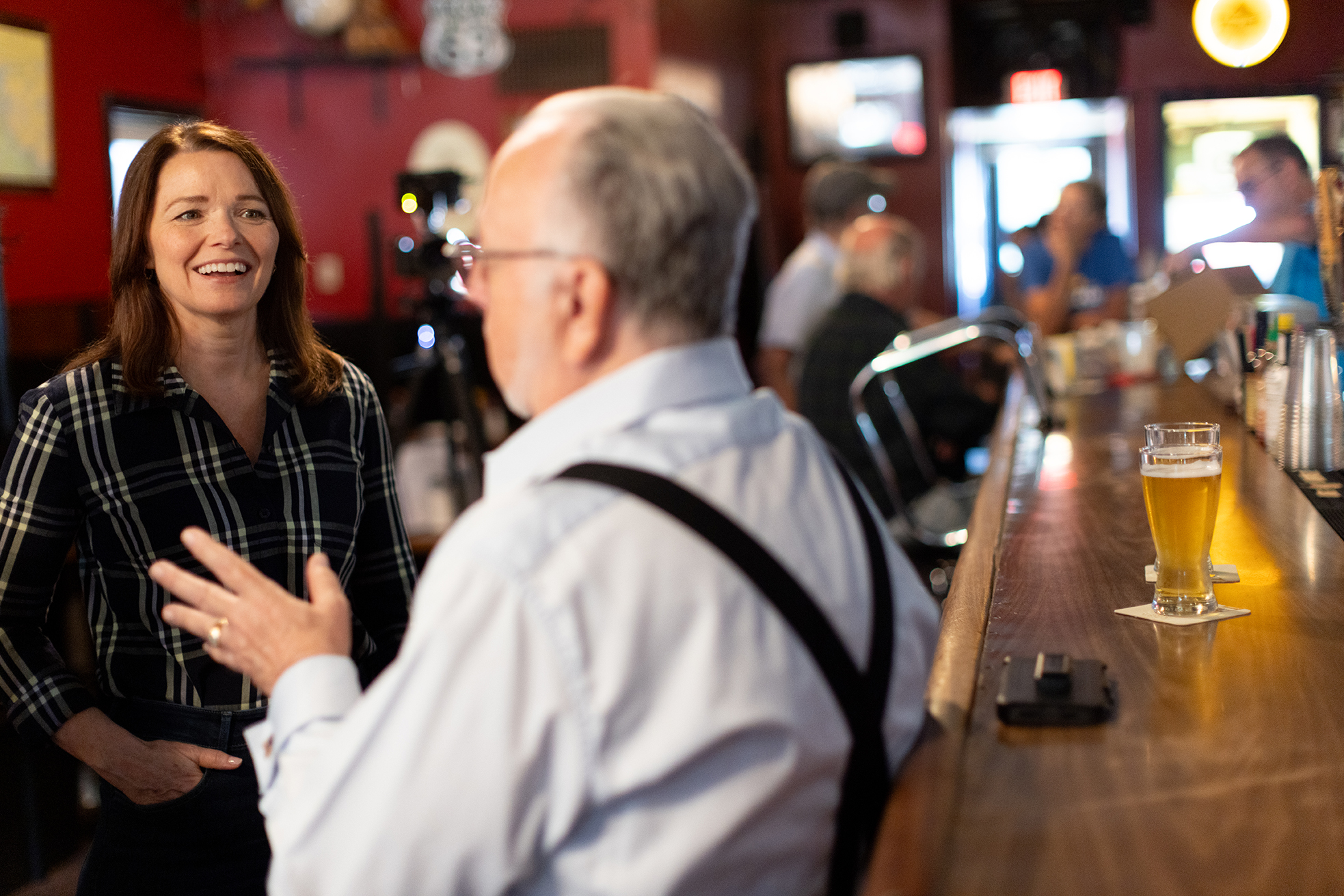 Christina Bohannan in a plaid shirt smiling and gesturing as she talks with a man in suspenders at a bar, a glass of beer on the counter