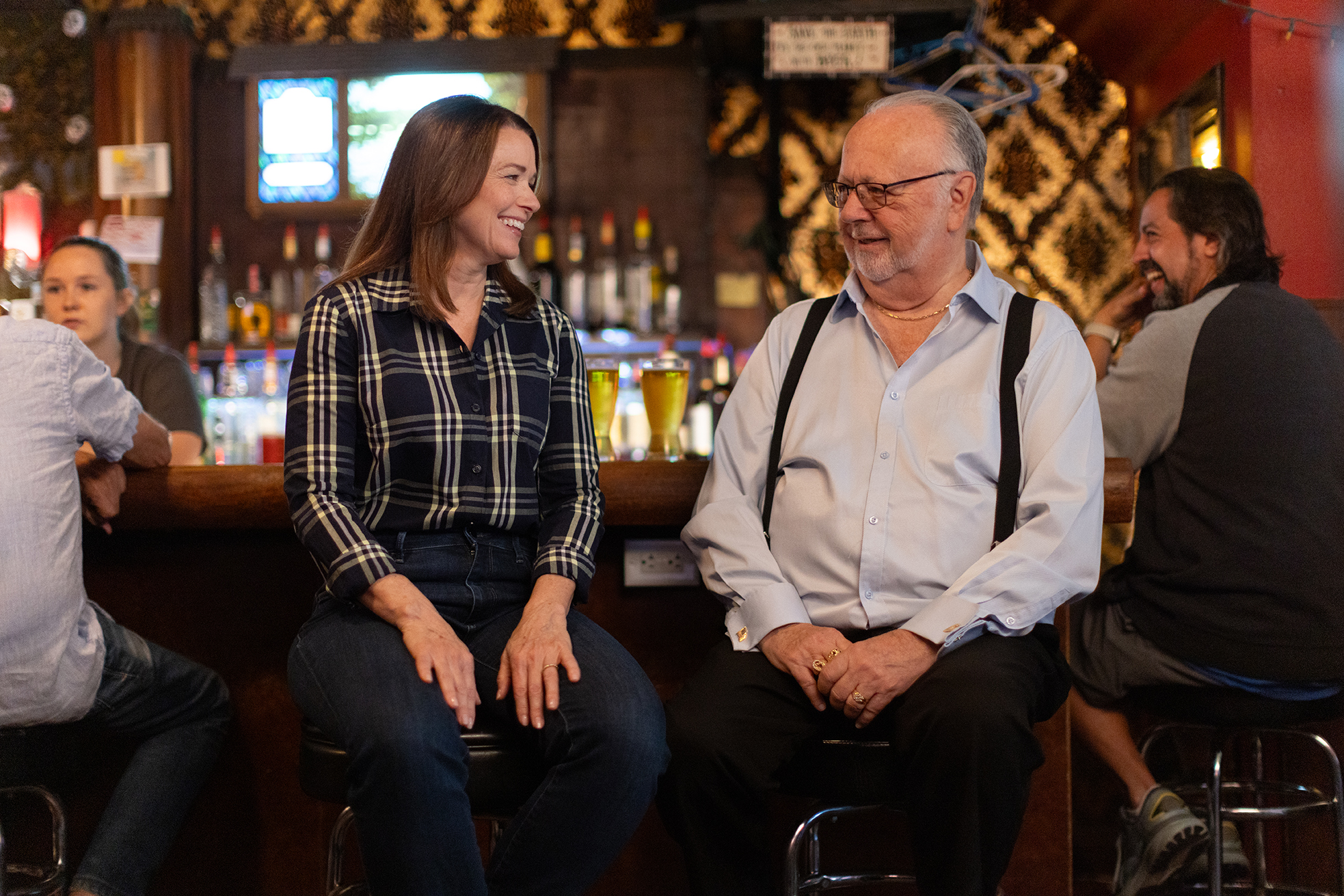 Christina Bohannan in a plaid shirt sitting and laughing with an older man in suspenders on bar stools at a bar