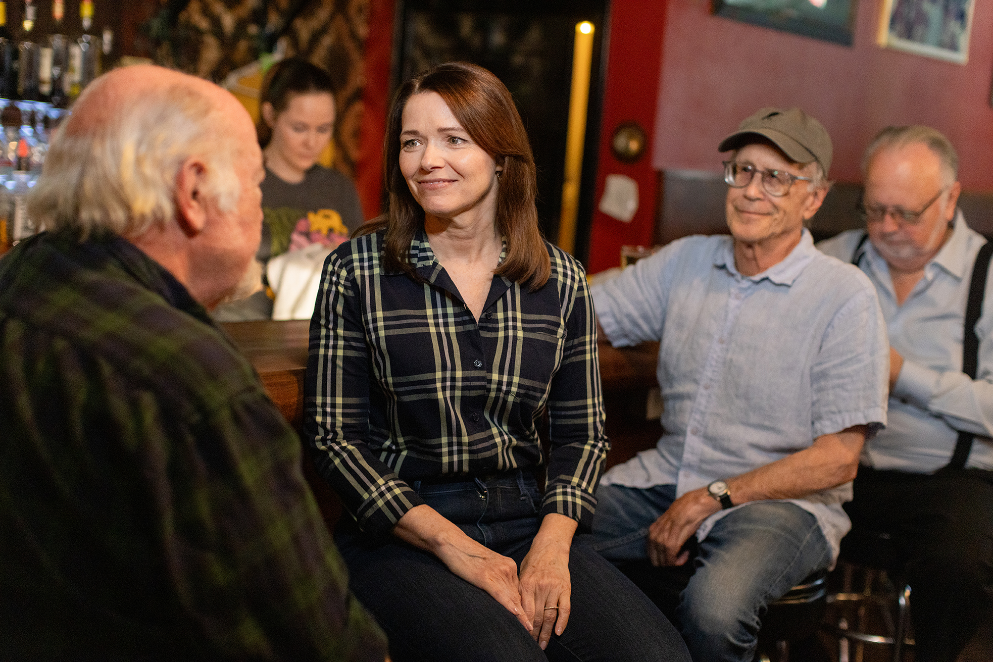 Christina Bohannan in a plaid shirt sitting and smiling among a group of people inside a dimly lit bar