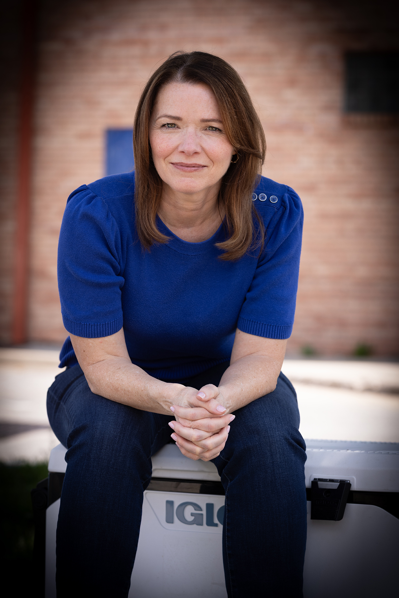 Christina Bohannan in a blue sweater sitting on a white cooler in front of a brick wall, hands clasped, smiling