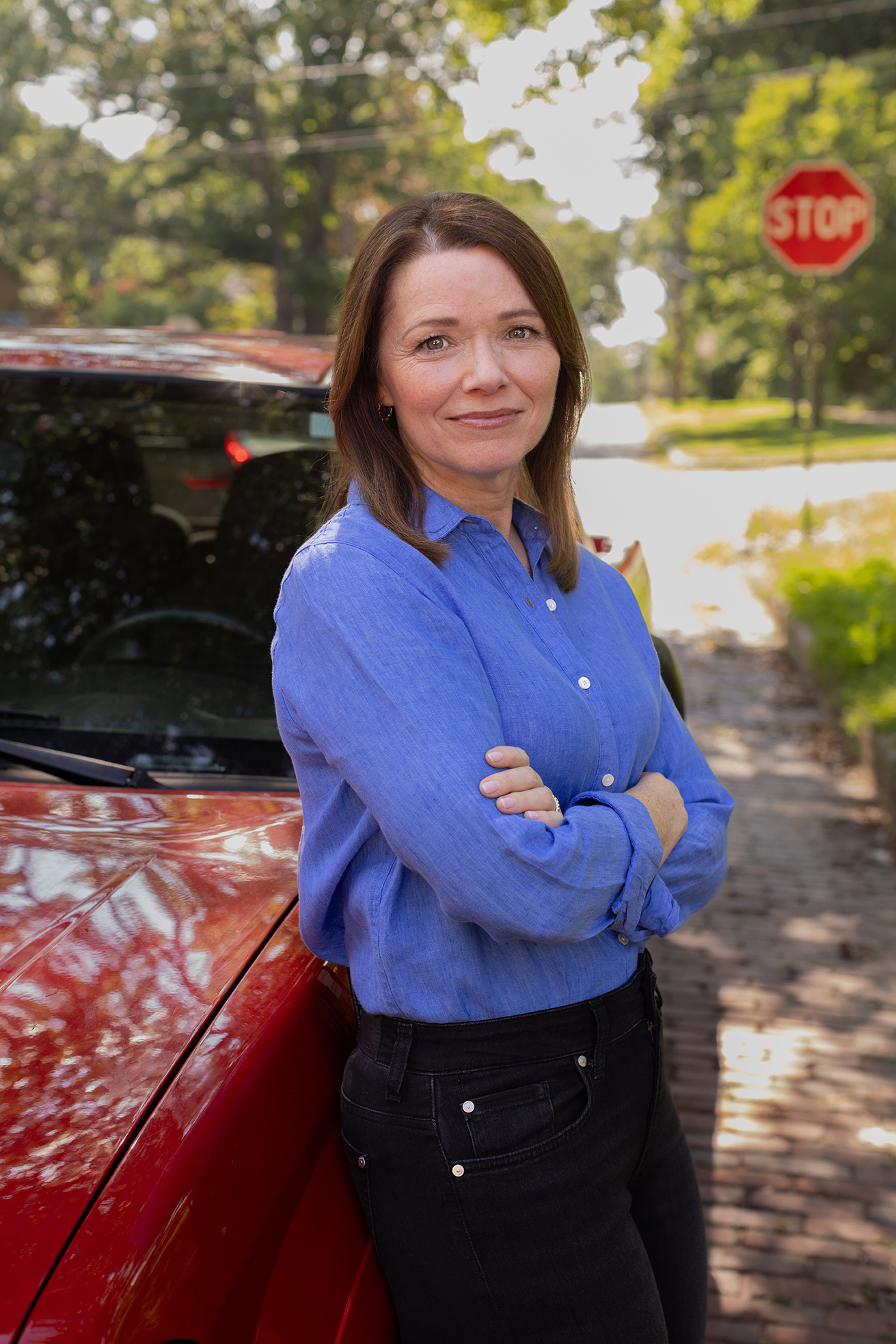 Christina Bohannan in a blue button-up shirt standing with arms crossed, leaning against a red car on a brick street near a stop sign