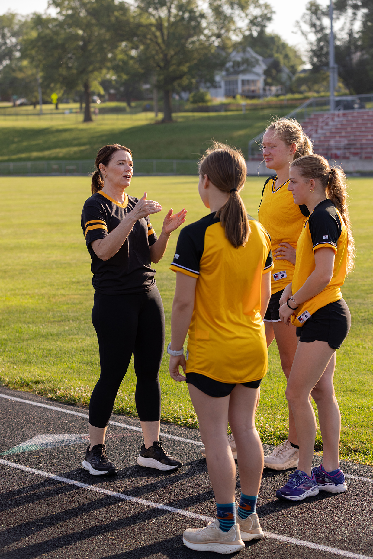 Christina Bohannan in a black and gold jersey gesturing while talking with three young female athletes in yellow and black uniforms on an outdoor track