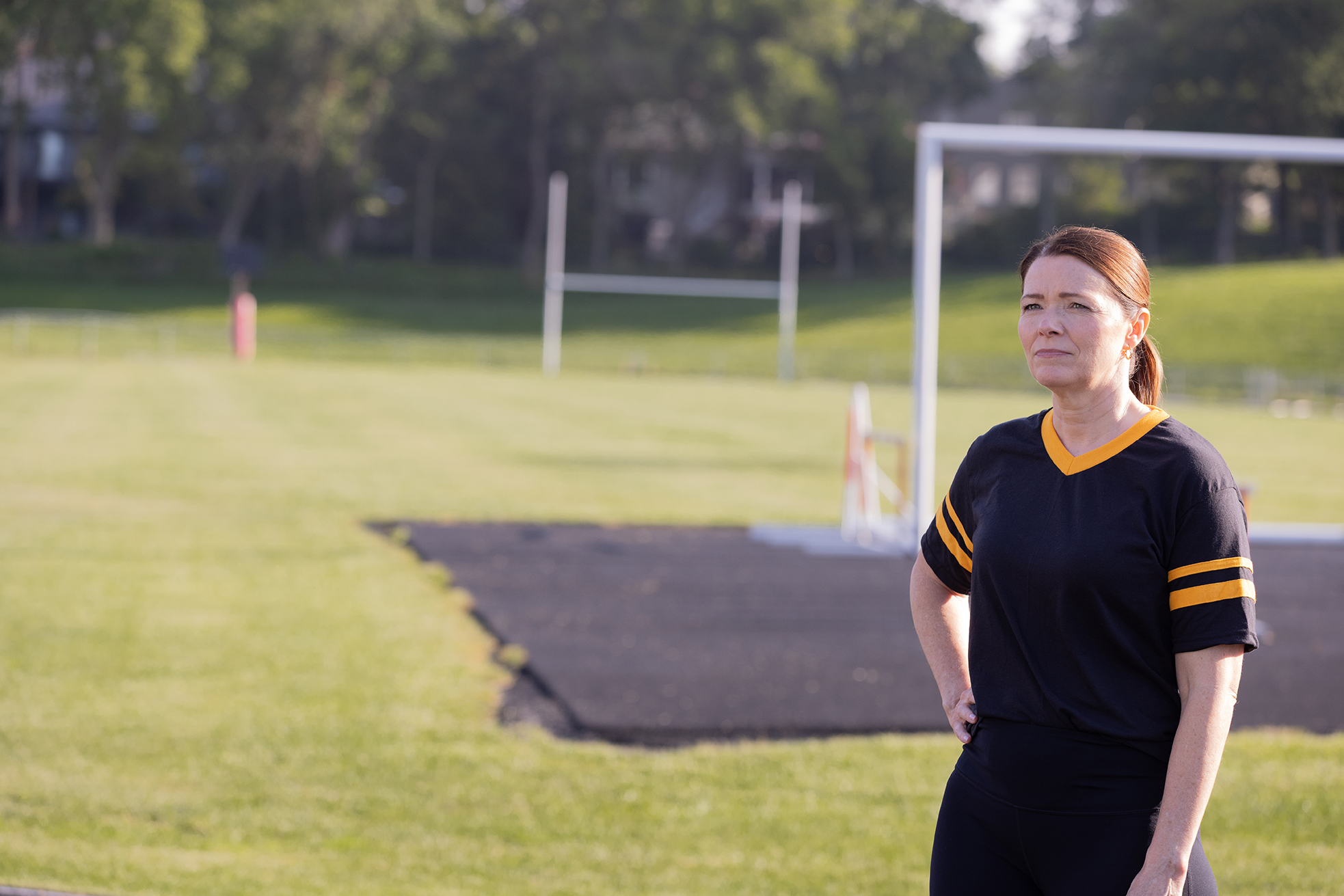 Christina Bohannan in a black and gold jersey standing on a grassy field with goalposts, looking into the distance