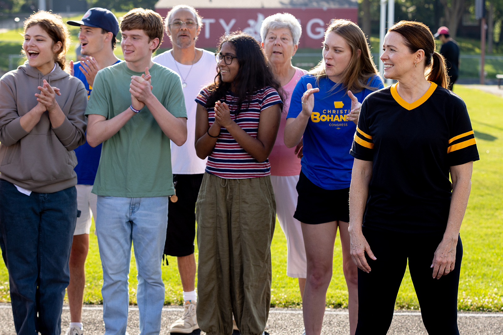 Christina Bohannan in a black and gold jersey standing beside a group of smiling, applauding young people outdoors, one wearing a blue campaign t-shirt
