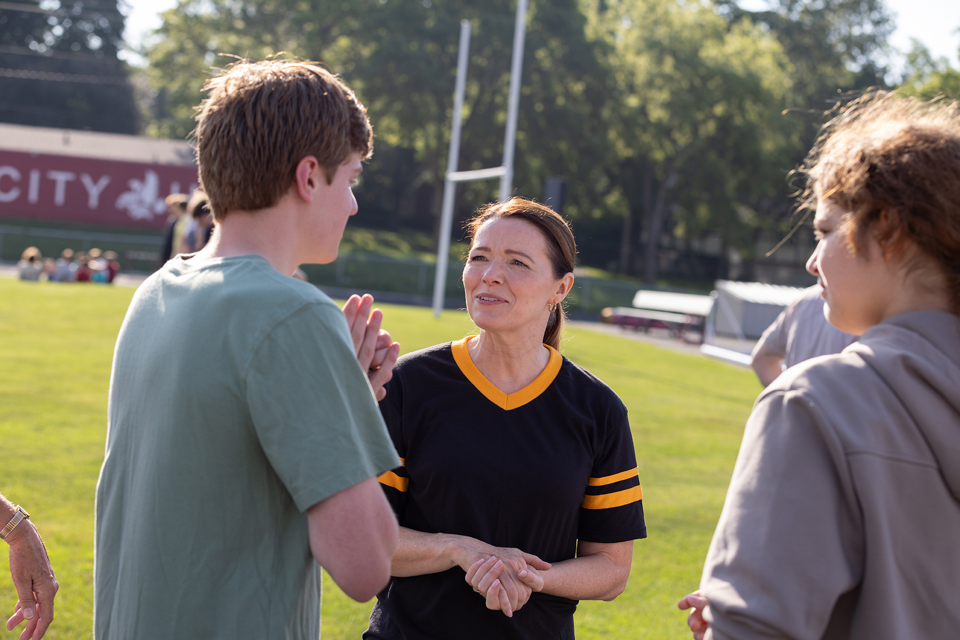 Christina Bohannan in a black and gold jersey talking with a teenage boy in a green t-shirt on a grassy sports field