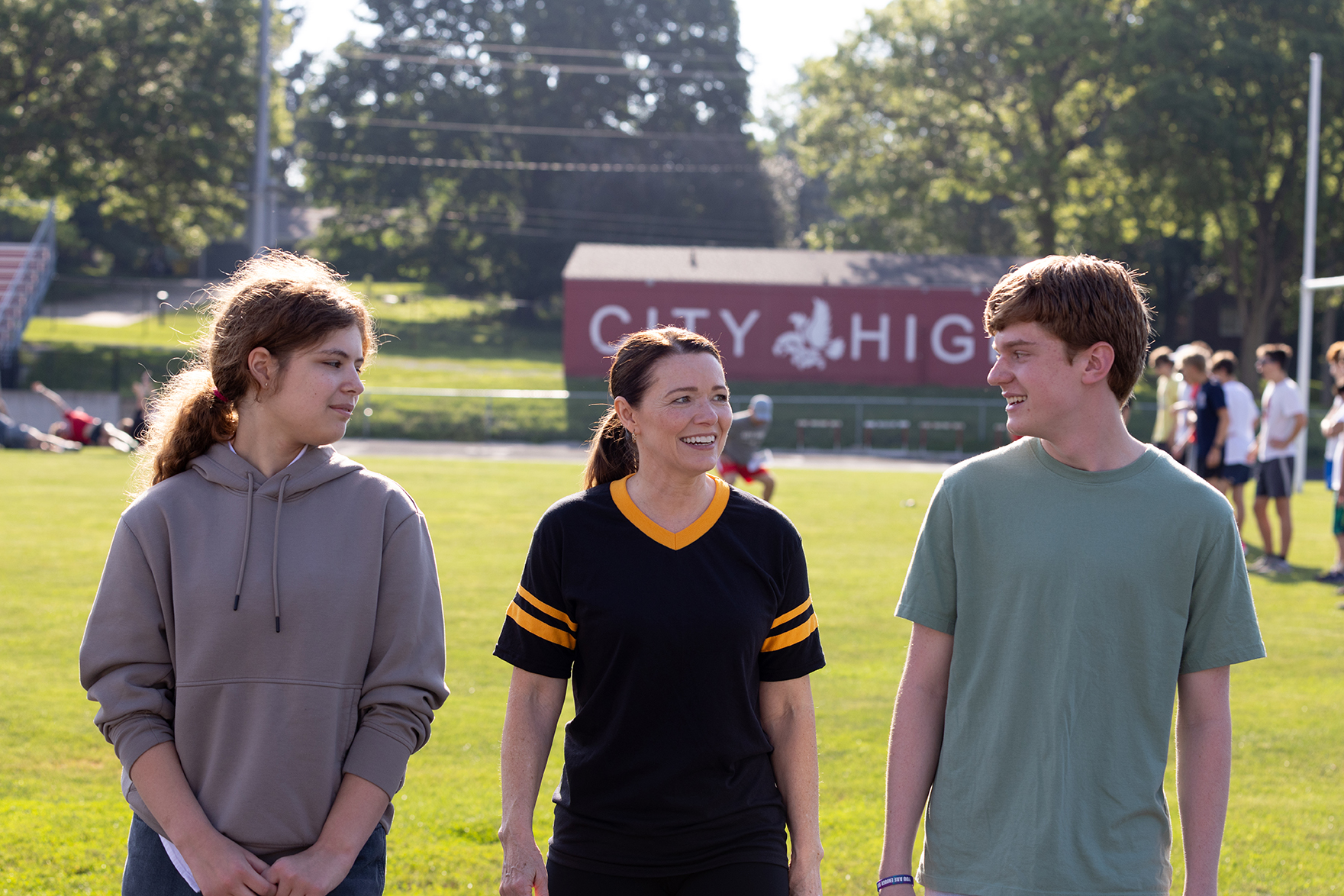 Christina Bohannan in a black and gold athletic jersey walking on a grassy field between a teenage girl in a gray hoodie and a teenage boy in a green t-shirt, with a City High sign behind them