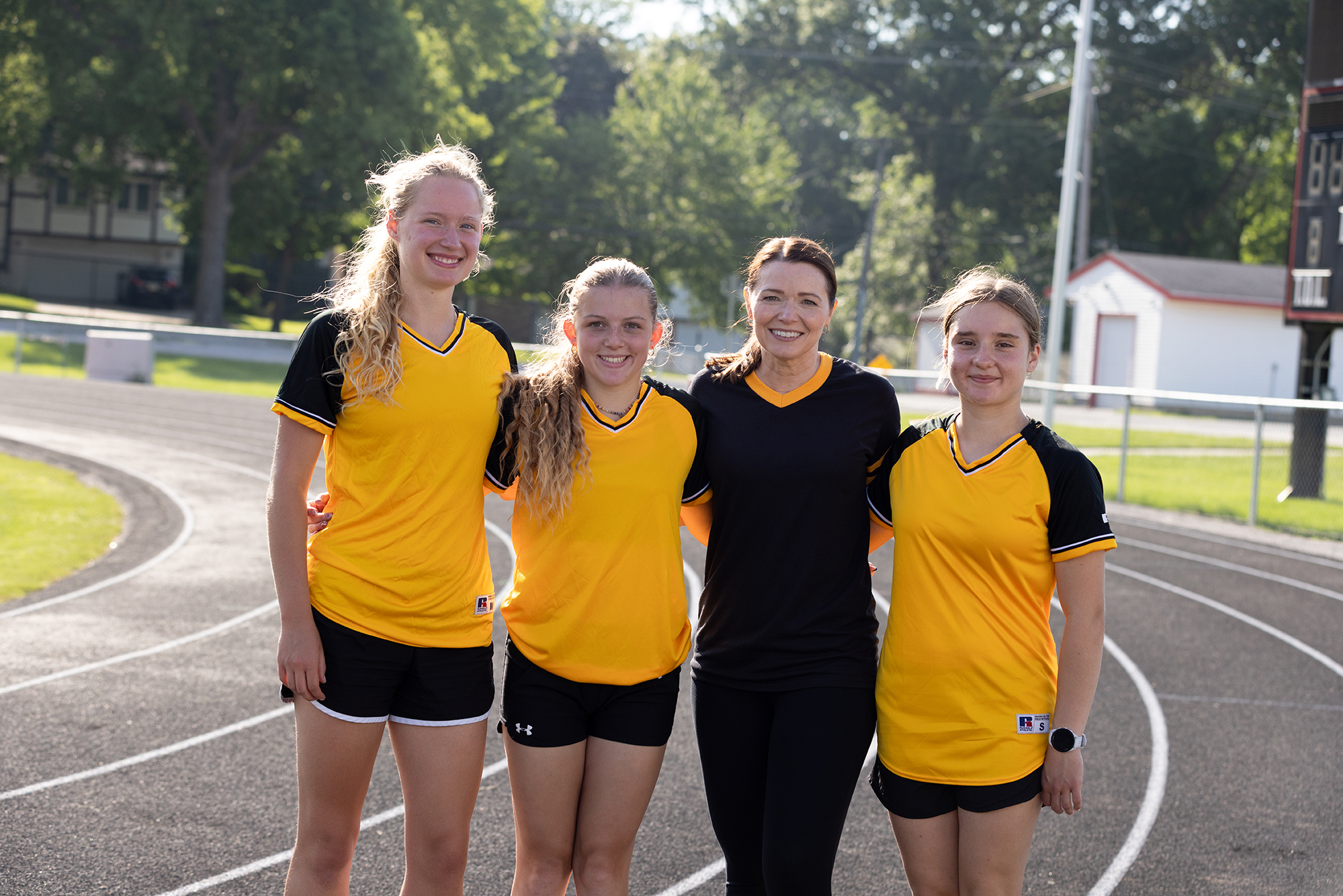 Christina Bohannan in a black athletic shirt with her arms around three young female athletes in yellow and black uniforms on an outdoor running track