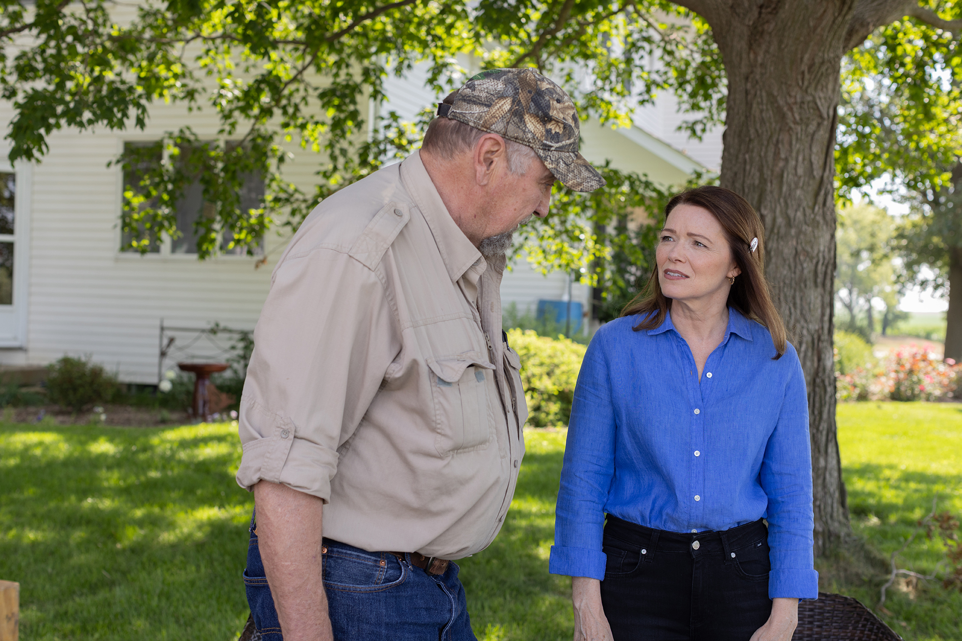 Christina Bohannan in a blue shirt talking with an older man in a camouflage cap and tan shirt in a yard in front of a white farmhouse