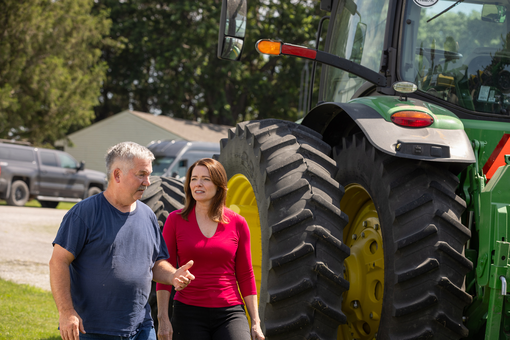 Christina Bohannan in a red top walking and talking with a man in a blue shirt beside a large green tractor on a farm
