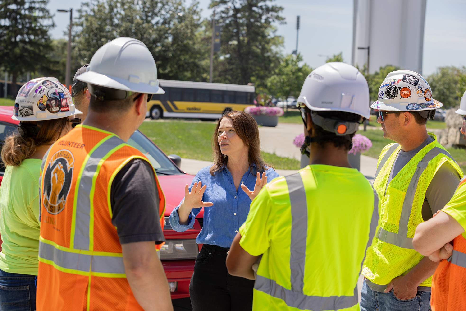 Christina Bohannan in a blue shirt gesturing while speaking with construction workers in hard hats and safety vests, with a school bus and red truck behind them