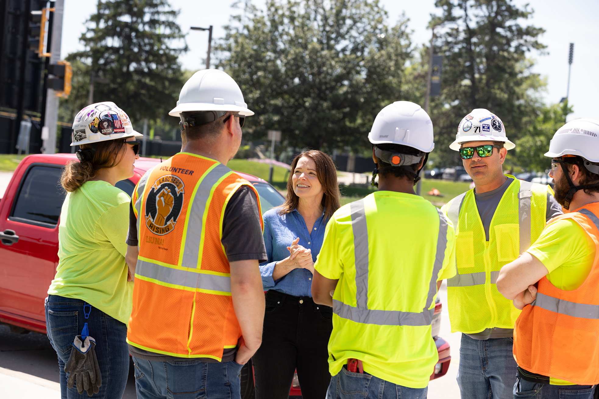 Christina Bohannan in a blue shirt talking with a group of construction workers wearing hard hats and high-visibility safety vests outdoors